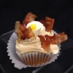 A bacon caramel cupcake topped with maple butter cream, bacon pieces, and a candy egg. Bacon & Eggs