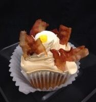 A bacon caramel cupcake topped with maple butter cream, bacon pieces, and a candy egg. Bacon & Eggs