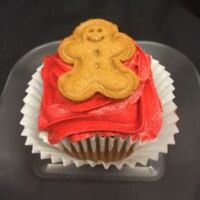 A gingerbread cupcake topped with red gingerbread cream cheese frosting and finished with a mini gingerbread man cookie. Gingerbread
