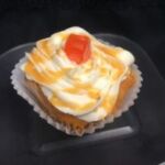 An orange white chip cupcake with orange ganache filing, topped with orange flavored cream cheese, an orange ganache drizzle and an orange candy piece. White Chocolate Orange
