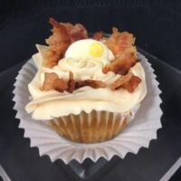 A bacon caramel cupcake topped with maple butter cream, bacon pieces, and a candy egg. Bacon & Eggs