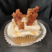 A cinnamon cupcake with bacon pieces, brown sugar cream cheese icing, and cinnamon sugar, topped with bacon pieces, and finished with a maple syrup drizzle. French Toast