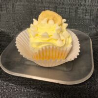 A banana cupcake with white ganache filling iced with banana buttercream and topped with a white ganache drizzle, chips, and a banana chip. White Chocolate Banana