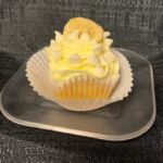 A banana cupcake with white ganache filling iced with banana buttercream and topped with a white ganache drizzle, chips, and a banana chip. White Chocolate Banana